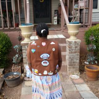 A person in a decorative vest and colorful skirt stands at the steps of a house, showcasing their unique style.