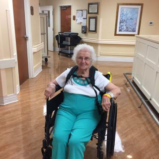 An elderly woman smiling while sitting in a wheelchair in a hallway.