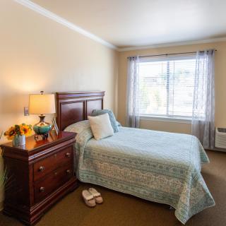 A cozy bedroom with a neatly made bed, nightstand, and a window letting in natural light.