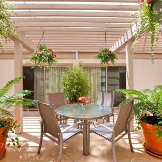 A cozy patio with a pergola, featuring a glass table and chairs, surrounded by lush greenery and hanging plants.