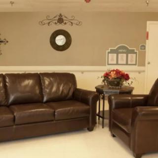 A cozy living room with a leather sofa, matching armchair, and a small coffee table adorned with flowers. The wall features decorative elements, including a clock.