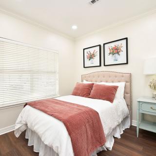 A cozy bedroom with a neatly made bed, accented with red pillows and a matching throw blanket. The room features two framed artworks above the headboard and a small nightstand with a lamp.