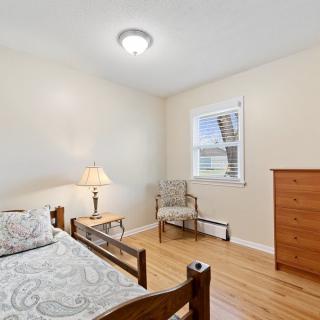 A simple bedroom with wooden furniture, featuring a bed, dresser, chair, and a table lamp beside the bed.