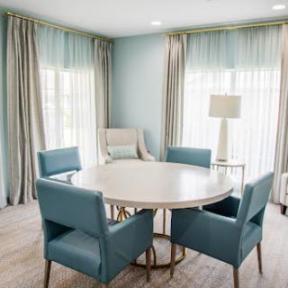 A modern dining area featuring a circular table surrounded by stylish blue chairs, illuminated by natural light filtering through sheer curtains.