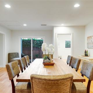 A modern dining room with a large wooden table and upholstered chairs, featuring minimalist decor and bright lighting.