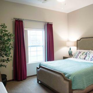 A cozy bedroom featuring a bed with a light turquoise blanket, burgundy curtains framing a window, and a bedside lamp next to a potted plant.