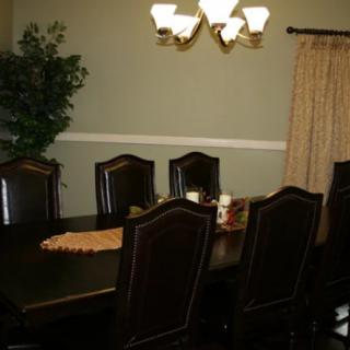 A dining room with a large table surrounded by chairs, a chandelier overhead, and decorative elements like a plant and curtains.