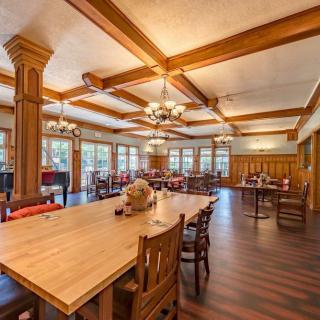 A spacious and warmly lit dining area features wooden furniture, large tables, chandeliers, and ample windows.