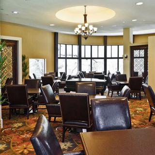 A well-furnished dining area with multiple tables and chairs, illuminated by a chandelier, featuring large windows and a colorful carpet.