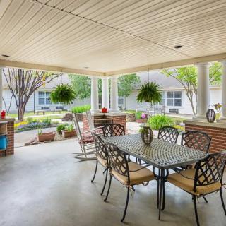 A spacious covered patio with a dining table and chairs, surrounded by a garden and potted plants.