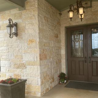 A cozy front porch with a stone wall and a wooden door, featuring lantern-style lighting and a planter.