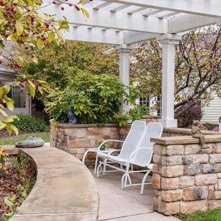 A cozy patio with a white pergola and chairs surrounded by lush greenery and stone walls.
