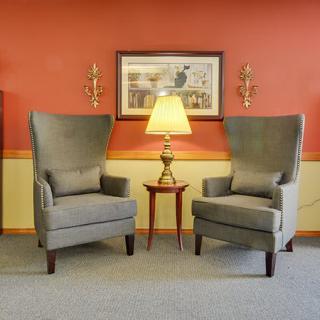A cozy living room setup featuring two grey armchairs, a round wooden table with a lamp, and wall decor.