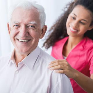 A smiling elderly man with a caregiver providing support.