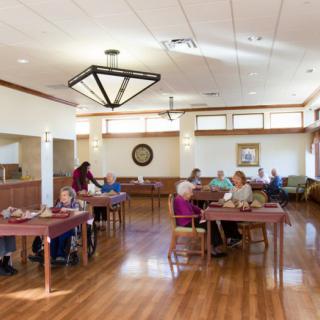 A communal dining area in a senior living facility with people seated at tables.