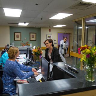 A busy hospital reception area with staff and visitors interacting at the front desk.