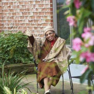 Elderly woman sitting outdoors in a garden setting, waving with a smile.