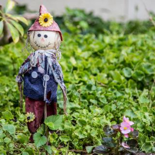 A small scarecrow stands in a lush garden surrounded by greenery and flowers.