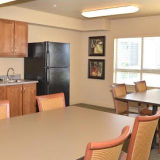 A modern kitchen and dining area with a refrigerator, table, and chairs, illuminated by natural light from the windows.