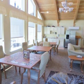 A bright and airy dining area with large windows, wooden ceiling, and a kitchen in the background.