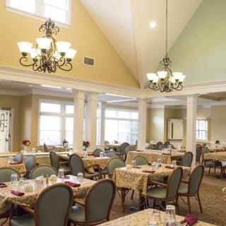 A spacious dining area with neatly arranged tables and chairs, illuminated by elegant chandeliers and natural light through large windows.