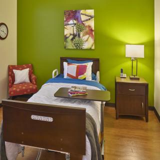 A cozy bedroom with a green accent wall, featuring a single bed, colorful artwork, and a warm lamp on the nightstand.