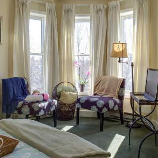 A cozy living room with large windows, two chairs, a floor lamp, and soft curtains.