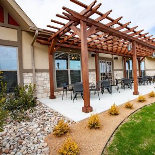 A modern patio with a wooden pergola and outdoor seating, surrounded by landscaped greenery.