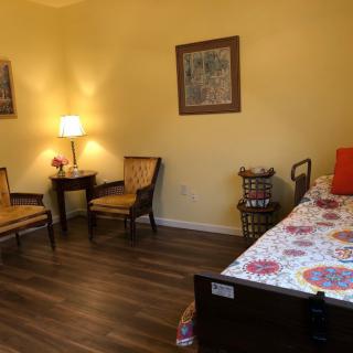 A cozy bedroom with wooden flooring, featuring a bed, two chairs, a table with a lamp, and colorful decor.