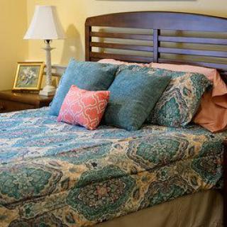A cozy bedroom scene with a neatly made bed featuring patterned bedding, multiple pillows, a nightstand with a lamp, and a picture frame.