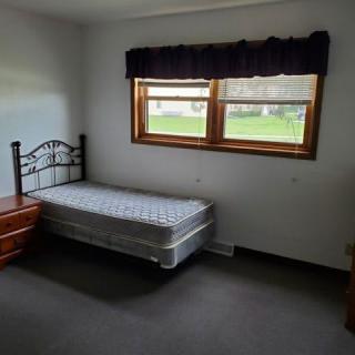 A minimalist bedroom featuring a single bed with a mattress, positioned beside a window.