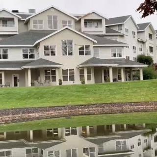 A large residential building with multiple stories is reflected in a pond, surrounded by a well-maintained lawn.
