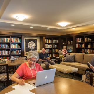 People reading and working in a cozy library setting with bookshelves and comfortable seating.