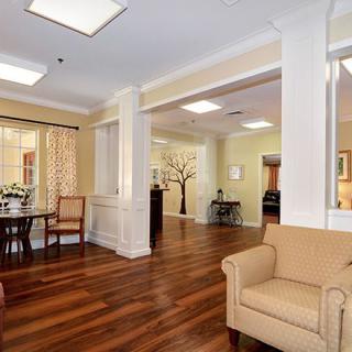 A well-lit living room with wooden flooring, featuring a comfortable sofa and a dining area in the background.