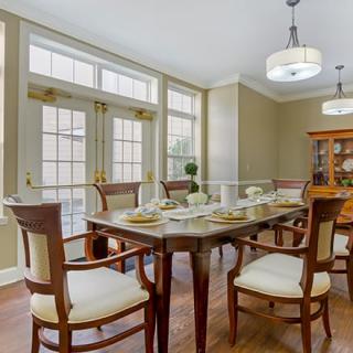 A well-lit dining room with a wooden table set and upholstered chairs, featuring large windows and elegant lighting.