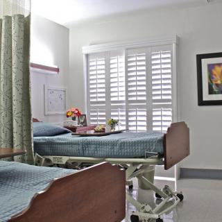 A hospital room with two patient beds, a dividing curtain, and a bright window with shutters.