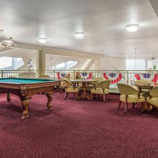 A spacious billiards room with a pool table and seating area, featuring red carpet and bright lighting.