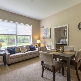 A cozy living room and dining area with a large window, featuring a comfortable sofa, dining table and chairs, and decorative elements including a lamp, artwork, and clock.