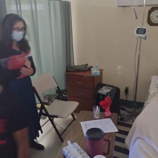 A caregiver in a mask interacts with a patient in a hospital room.