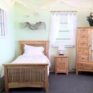 A cozy bedroom with light wooden furniture, including a single bed, nightstand, and dresser against light green walls.