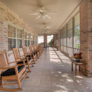 A spacious porch with several wooden rocking chairs lined up against a brick wall, featuring ceiling fans and bright natural light.