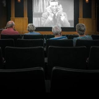 A group of elderly individuals watching a black and white film in a cozy cinema setting.