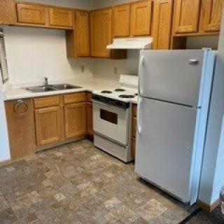 A small apartment kitchen with wooden cabinets, a refrigerator, stove, and a sink.