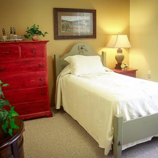 A cozy bedroom with a single bed, red dresser, and soft lighting from a table lamp.