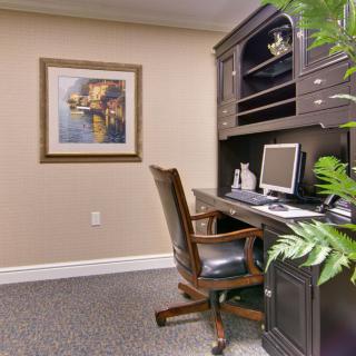 A cozy home office with a wooden desk, computer, and comfortable chair, accented by plants and artwork on the wall.