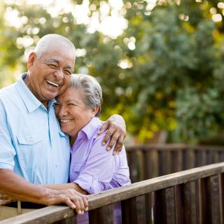 An elderly couple embracing while standing on a wooden bridge in a park.
