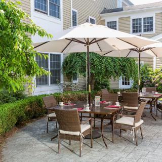A cozy outdoor patio area with a table and chairs under a large umbrella, surrounded by greenery and the exterior of a building.