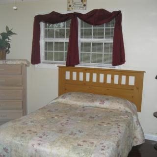 A cozy bedroom with a single bed, wooden headboard, and floral bedspread, accompanied by a dresser and windows with red curtains.