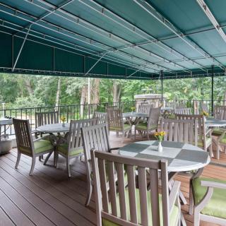 A peaceful outdoor patio with wooden tables and chairs under a green canopy, surrounded by lush greenery.