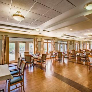 A spacious dining area with wooden floors, arranged tables and chairs, and large windows providing natural light.